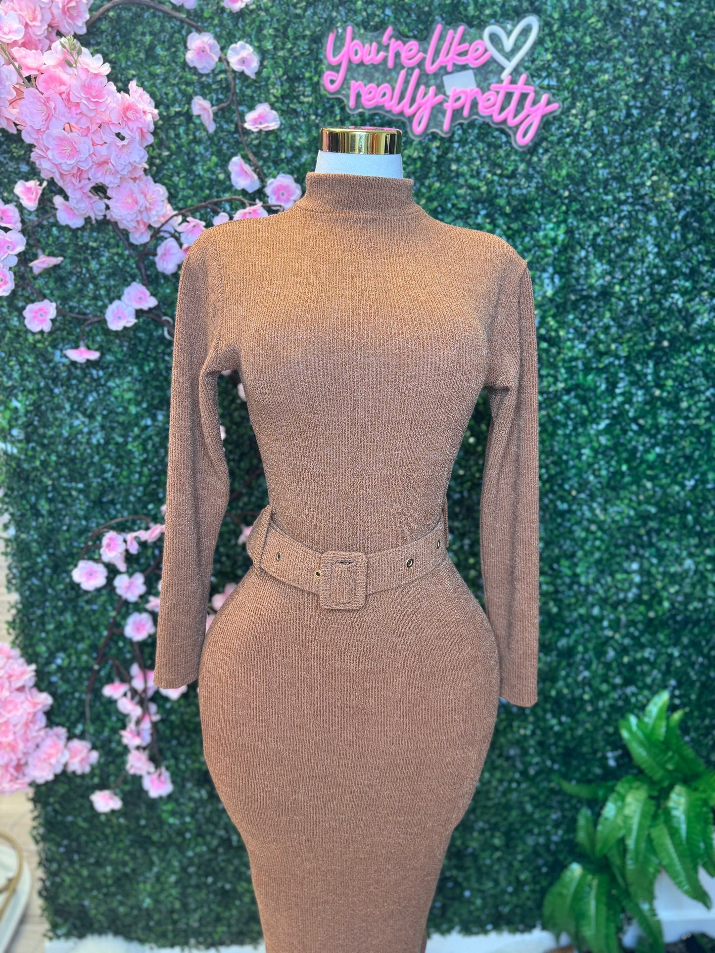 Mannequin wearing a brown ribbed dress with a belt against a decorative background with pink flowers and greenery.