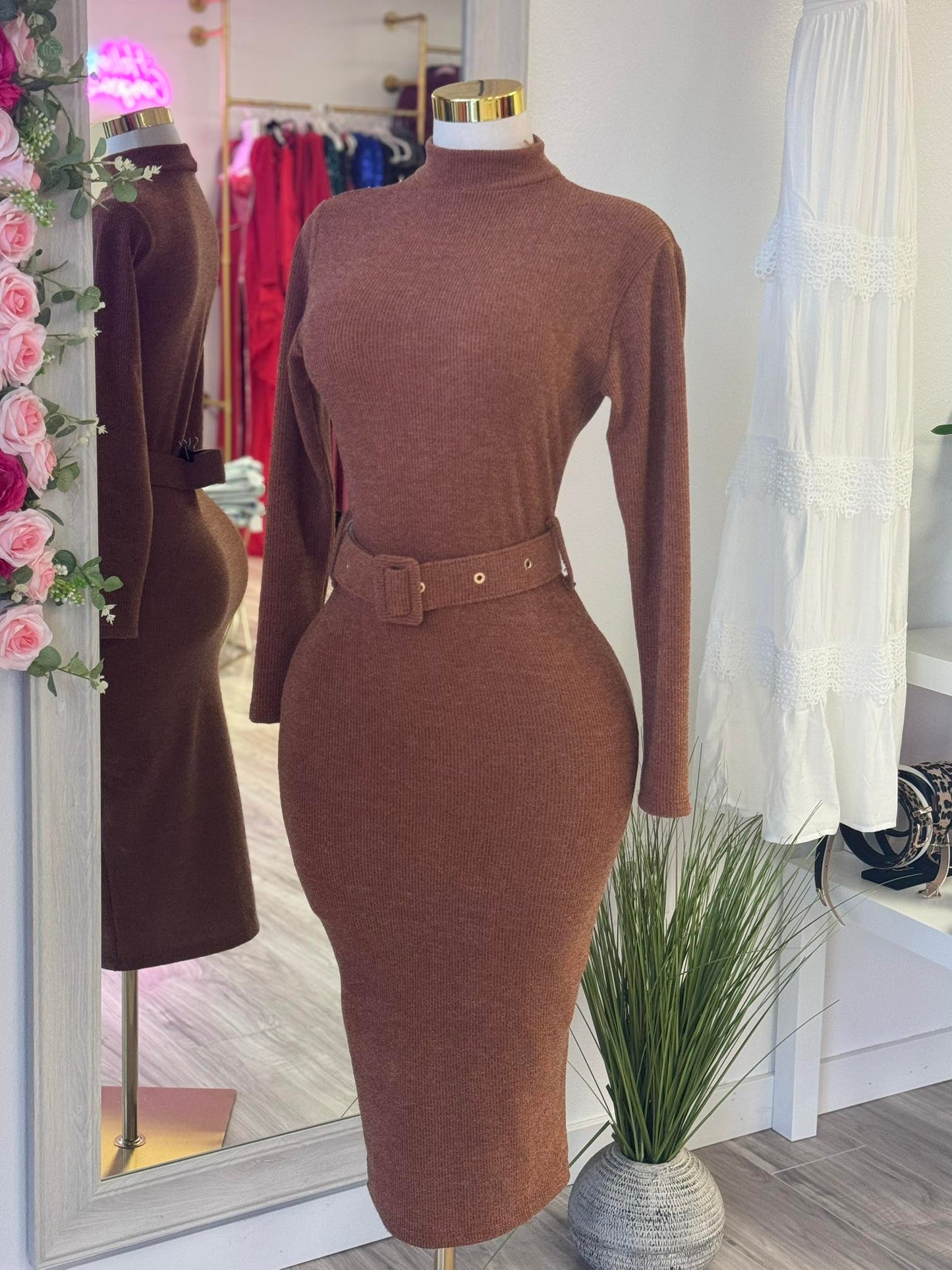 Brown fitted dress on a mannequin in a store setting with flowers and clothing in the background.