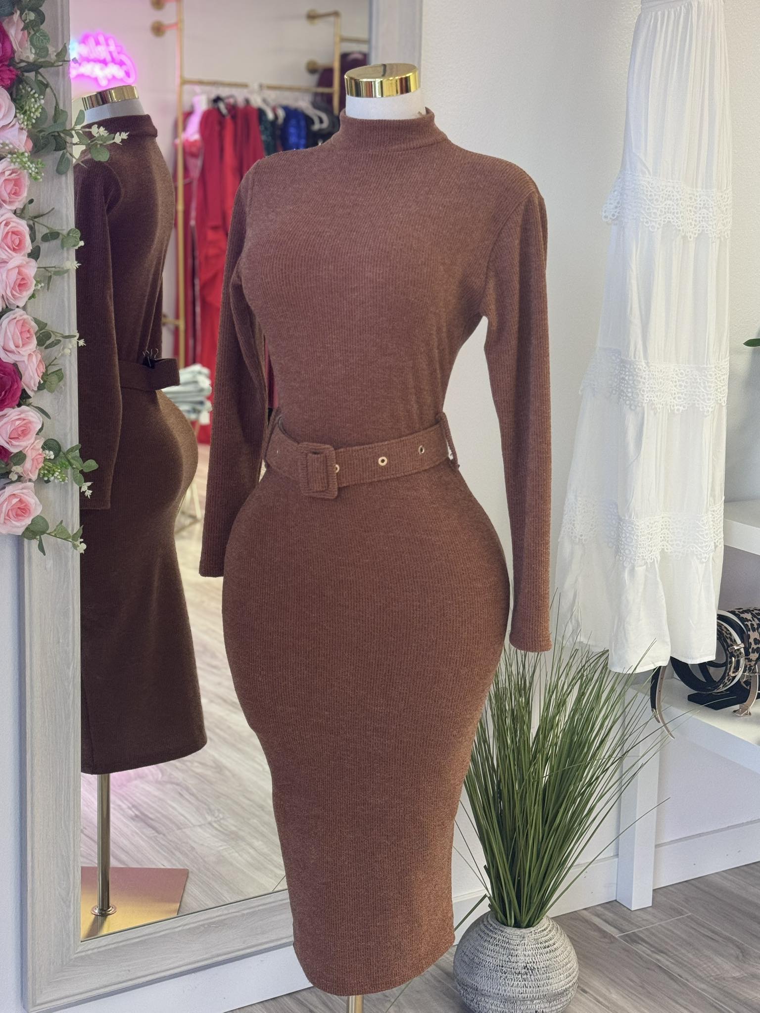 Brown fitted dress on a mannequin in a store setting with flowers and clothing in the background.